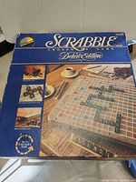 Top view of Scrabble Deluxe Edition box showing game name, imagery of board and game features, and visible shelf wear.