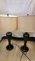Pair of side table lamps with beige drum shades and dark bases standing on floor, cords visible