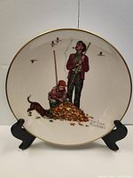 Front view of vintage Norman Rockwell 'Pensive Pals' decorative plate showing two boys raking leaves with dog and birds, signed by Norman Rockwell. Plate on black stand.