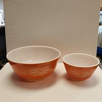 Photo of two orange Pyrex mixing bowls with Harvest Wheat pattern, showing larger and smaller bowl side by side on white background.