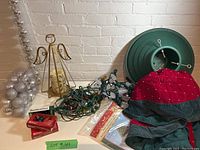 Photo showing assorted Christmas decorations including angel candle holder with candle, bag of silver balls, Christmas tree stand, bundles of string lights, and red tree skirt.