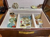 Opened wooden jewelry box with assorted costume jewelry brooches and kilt pins arranged in three velvet-lined compartments