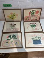 Overview of all six framed folk art floral paintings arranged on white table against white brick wall