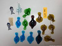 Top-down view of 14 glass and ceramic fish figurines in assorted colors arranged on white surface with orange label showing lot number.
