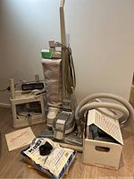 Photo showing Kirby Generation 3 vacuum with cloth bag, flexible hose, separate carpet shampoo attachments in a plastic container, spare vacuum bags, and user manual on floor against wall.