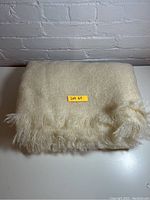 Folded cream coloured mohair throw with fringed edges displayed on white surface against white brick wall.