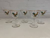 Set of five small clear stemmed glasses with colorful rooster images and gold rims, arranged in a row on a white surface.