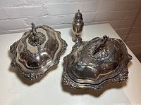 Two silver plated chafing dishes with ornate handles and a decorative silver plated vessel, all arranged on a white surface against a white brick wall background.