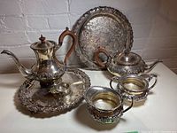 Silver tea and coffee set displayed with two round trays, cream and sugar containers, and a tea strainer. Items show ornate engraving and wooden handles.
