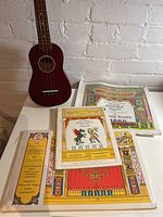 Full lot including Dgas Ukulele and three Pollocks children's toy theatre sets shown flat on a table, ukulele reddish wood body, theatre sets with colorful prints.