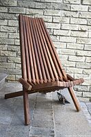 Front angled view of Tamarack folding chair showing slatted acacia wood seat and backrest with cinnamon finish against brick background.