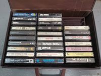 Top view of 27 cassette tapes arranged in a brown carrying case, various artists including Led Zeppelin, Boston, KISS, AC/DC, The Doors