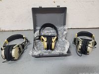 Three beige and black Fostex over-ear headphones with one set inside an open gray hard plastic carrying case lined with bubble wrap.