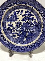 Blue and white Crown Pottery plate showing a Chinese mosaic scene with figures crossing a bridge, a boat on the river, and birds near a temple entrance, displayed on a stand.