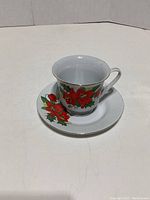 Teacup placed on saucer showing front floral design with poinsettias and holly leaves and gold trim.