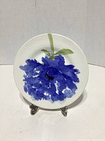Front view of the white ceramic plate with blue flower and green stem design on stand against plain background.