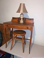 Antique wooden desk with raised back compartments, matching round wooden stool, ivory table lamp with beige shade on top of desk in a room.