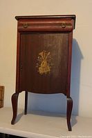 Front view of antique cherry wood music cabinet showing gold painted musical instruments and sheet music on front door, with ring pull handles on upper drawer