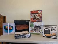 Photo showing all items in lot on table including waffle maker, pasta/rice cooker, 16-inch roasting pan, mixing bowls, and silverware organizer, all boxed and new.