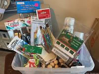 Photo showing a bin of various craft supplies including wooden beads, multiple wooden ornaments such as snowman and tree shapes, packs of beads and sequins, and multiple sets of paint brushes and paints.