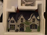 Porcelain house figurine depicts Brownlow House from Oliver Twist series with blue roof, stone-textured walls, ivy accents, name plaque, chimney stacks, and detailed windows and door.