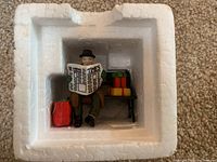Figurine of a man sitting on a bench reading a newspaper with packages beside and bags at feet inside original white foam packaging.