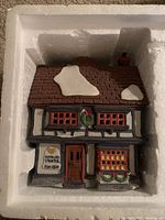 Color photo of Dept 56 Tutbury Printers ceramic building inside styrofoam packaging, showing painted snowy roof and holiday wreath decorations.
