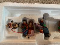 Figurines in original white foam packaging showing a child holding a cow on a leash, seated man and woman carolers in winter clothing.