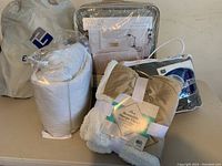 Photo shows white rolled blanket, beige reversible sherpa fleece blanket, beige mattress pad, folded sheets all grouped together.