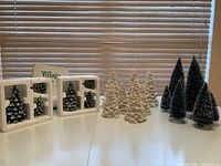 Image showing four sets of Christmas trees including Department 56 boxed village evergreen trees, white porcelain trees, and green craft trees arranged on a table.