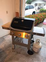 Weber Spirit 2 gas grill with three burners, black hood with built-in thermometer, stainless steel front and side shelves, propane tank attached, placed inside garage.