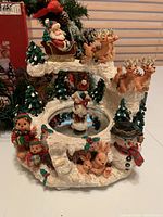 Christmas music box showing Santa Claus in sleigh led by reindeer above a snowy circular base with trees, rabbits, teddy bears, and a snowman, along with two separate magnetic skating figurines.