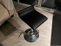 Image shows one of two black leather seat barstools with chrome bases and footrests, placed on wooden floor near window at night.