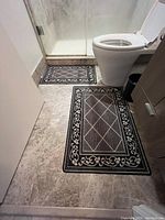 Photo showing two rectangular bathroom mats in brown and black placed on a bathroom floor near a toilet and shower.