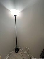 Photo of a tall black floor lamp with a round base and a white frosted glass shade lit to show ambient lighting in a corner of a room.