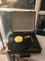 Photo of the Crosley record player inside a suitcase-style wooden case, lid open, showing the turntable with a vinyl record on it, the tonearm, and control knobs.