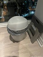 Small white or off-white kitchen garbage can with dome lid. Positioned inside with a trash bag. Next to a small step stool, placed on wood flooring near a window overlooking city lights at night.