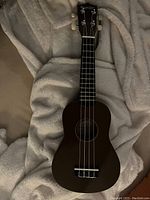 Brown ukulele lying on a soft beige blanket, showing full front view with four strings, black fretboard, and cream-colored tuning pegs.