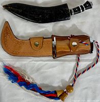 Top view of two decorative blades in their sheaths laid on white fabric. One sheath is black leather, the other light brown tooled leather with colorful tassels.