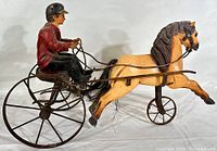 Side view of folk art harness racing sculpture showing horse and driver on sulky cart with metal wheels.