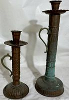 Front view of two asymmetrical red copper candlesticks with brass handles, showing the varied heights and oxidation on the taller one.