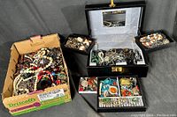 Wide shot showing jewelry box with open compartments revealing mixed costume jewelry and a cardboard box filled with more jewelry items including necklaces and bracelets.