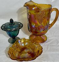 Three piece set with Indiana amber Harvest Grape Carnival glass pitcher, blue iridescent covered candy dish, and Loganberry amber iridescent bowl. Set displayed on white background showing detail and translucency.