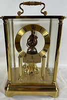 Front view of brass mantel clock showing transparent glass housing, circular dial with Roman numerals, and brass handle on top.