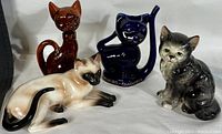Four vintage porcelain cat figurines displayed together on a white fabric background, showing variety in color and pose.