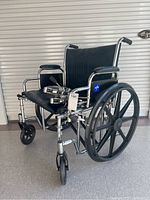 Front-angle view of chrome frame wheelchair with wide black vinyl seat, large rear wheels, and front caster wheels, showing footrests attached.
