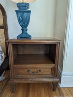 Front view of wooden bedside table with one drawer and open shelf, with blue lamp on top (lamp not included).
