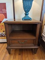Front view of Gibbard bedside table showing one open shelf and one drawer with brass handle, wood floor background, blue decorative lamp on top.