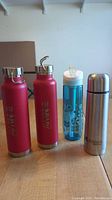 Front view of four travel bottles showing two pink aluminum water bottles, one blue plastic bottle, and one silver thermos