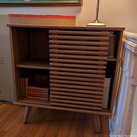 Front view of wooden small cabinet showing open shelving on left, sliding door partially covering right shelves, wood legs visible.
